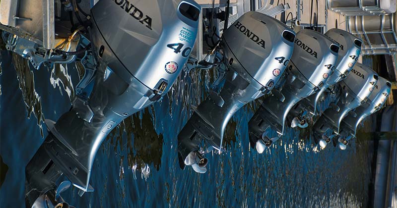 Zdjęcie przedstawia rząd kilku silników zaburtowych marki Honda, wyciągniętych ponad wodę z widocznymi śrubami