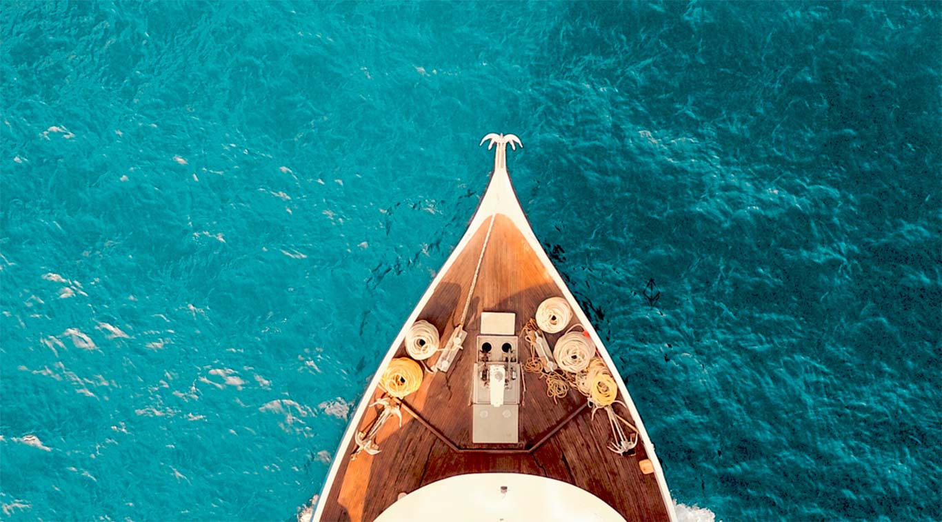 Widok prostopadle od góry z lotu ptaka na dziób białej łodzi motorowej z drewnianym pokładem, płynącej po turkusowych, egzotycznych wodach